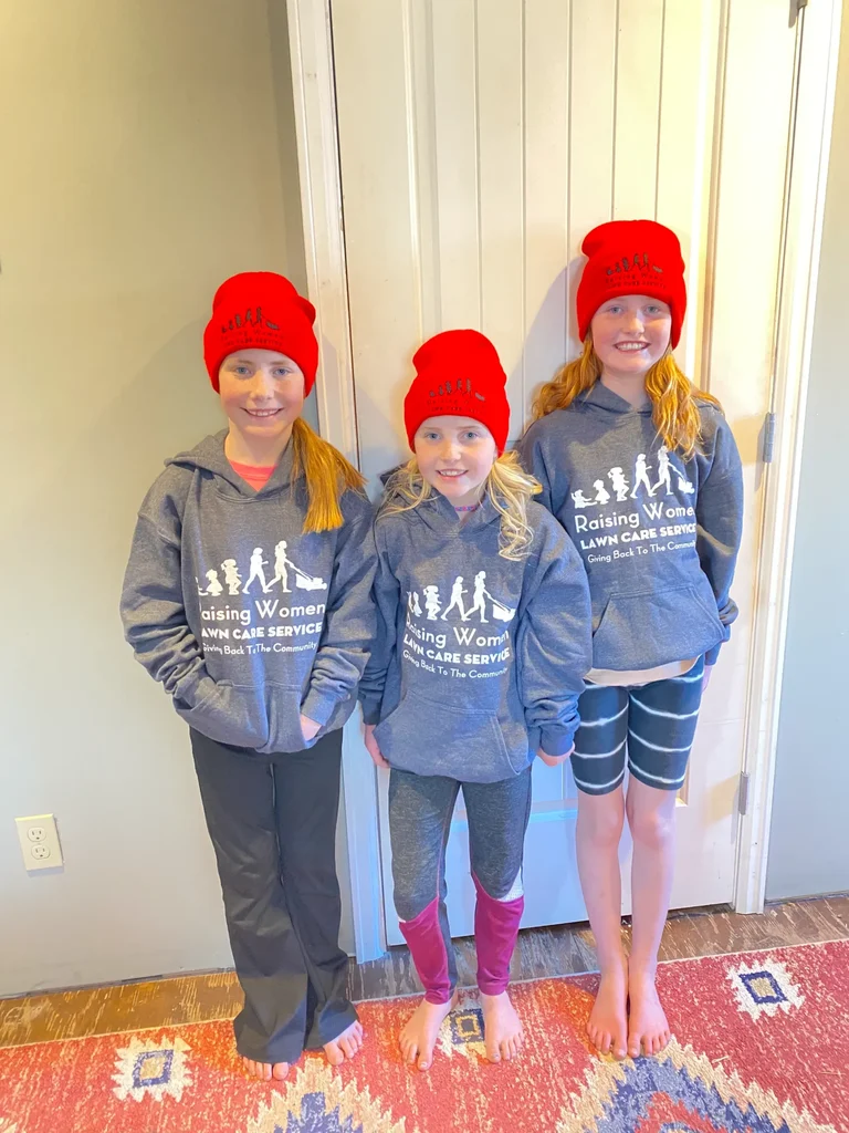 Three girls wearing Raising Women shirts and red winter hats